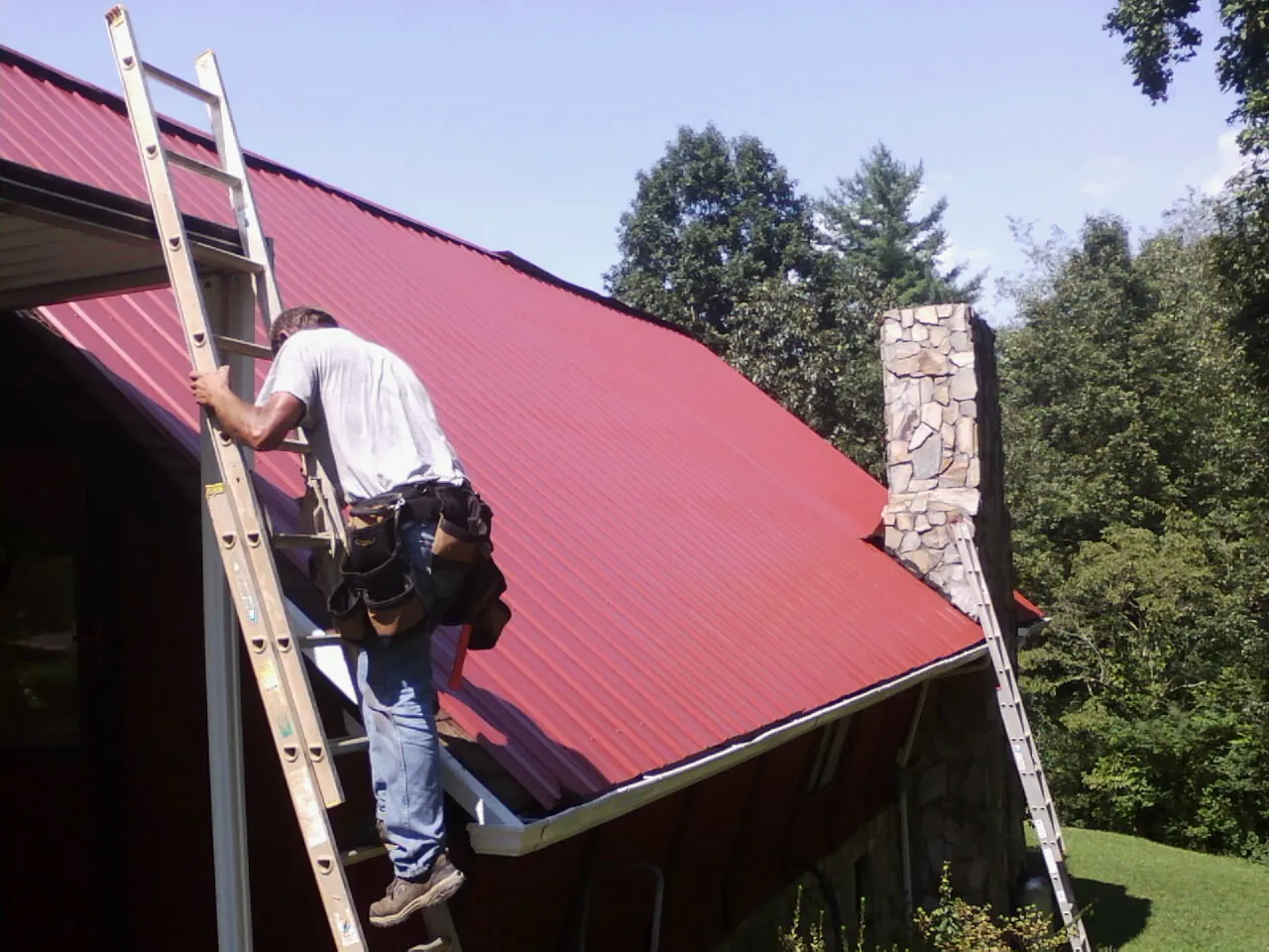Roofer on a ladder beside a red metal roof and stone chimney — Soffit & Fascia Repair by Roofing Brothers in Walkerton